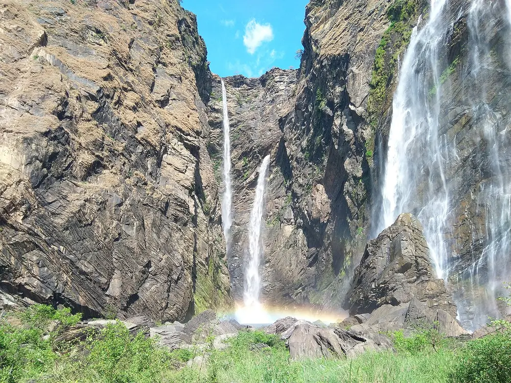 Thin streams of water falling down a towering grey cliff into a green gorge, with a small rainbow at the bottom.