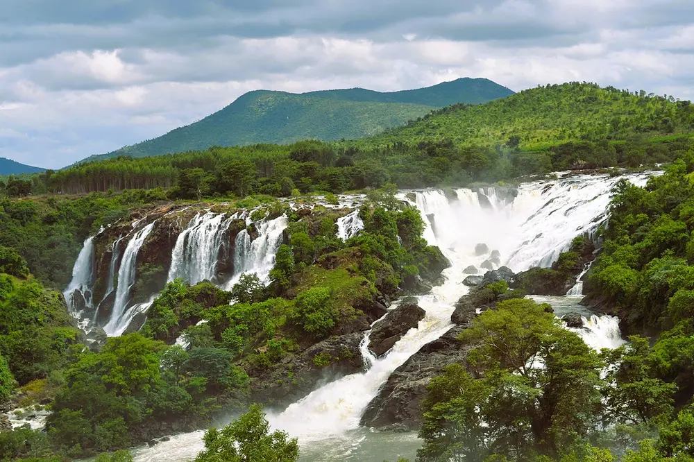 A wide waterfall spilling in many white streams over a rocky, forested ledge, with green hills behind.