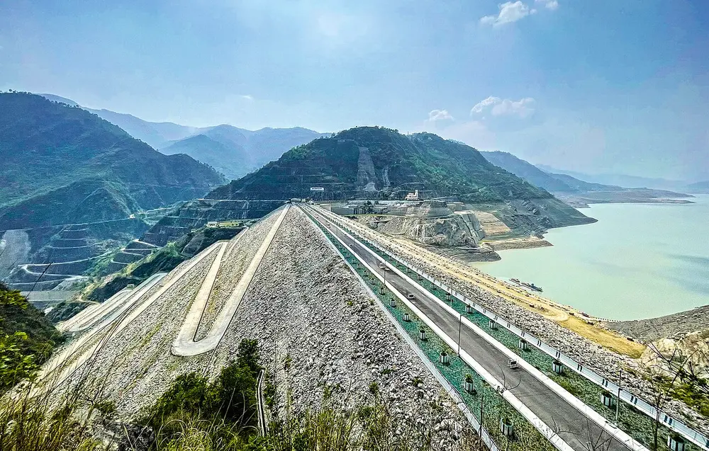 A huge sloping wall of rock and earth with a road zigzagging down it, holding back a lake among green mountains.
