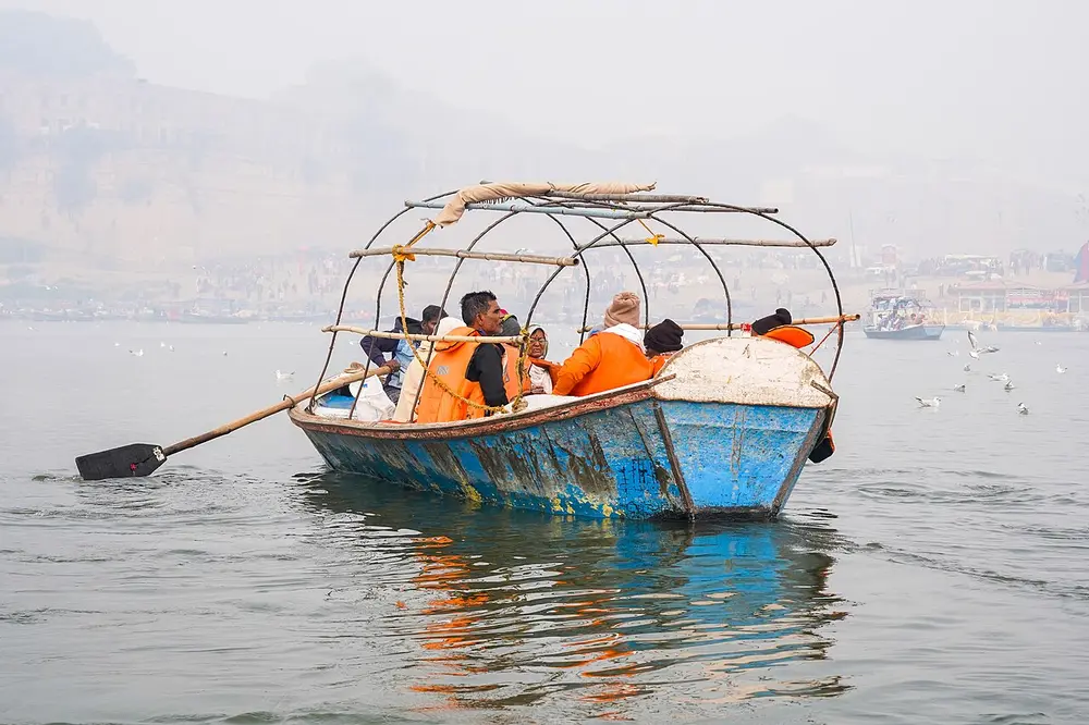 A blue wooden rowing boat with pilgrims in orange robes on hazy water, with gulls flying behind.
