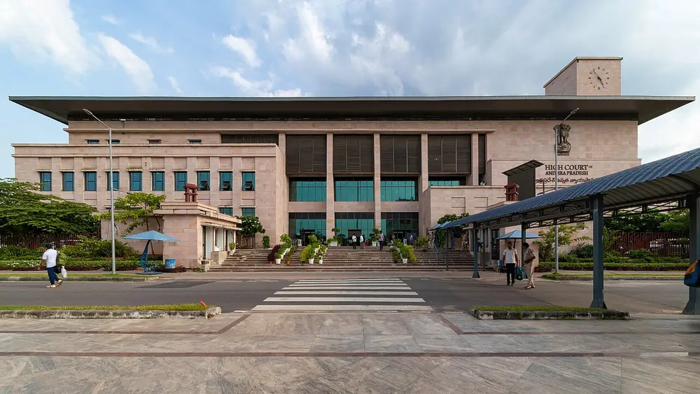 A long, pale pink stone building with a wide flat roof, tall windows and a clock tower at one corner, behind a pedestrian crossing, with a sign reading High Court of Andhra Pradesh.
