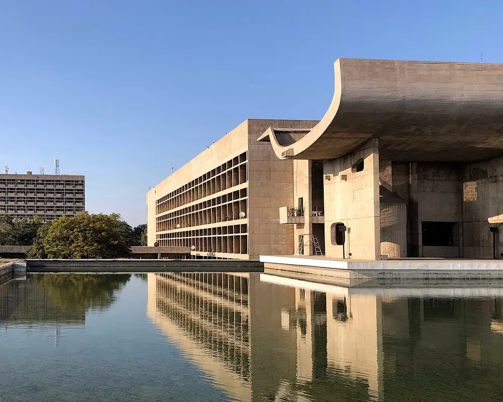 A concrete government building with a huge upturned curved roof over its entrance, reflected in a pool.