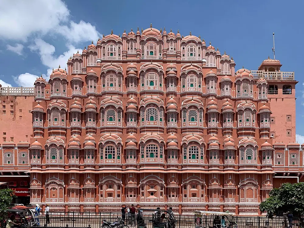 A tall pink sandstone palace front covered in hundreds of small latticed windows, under a blue sky.