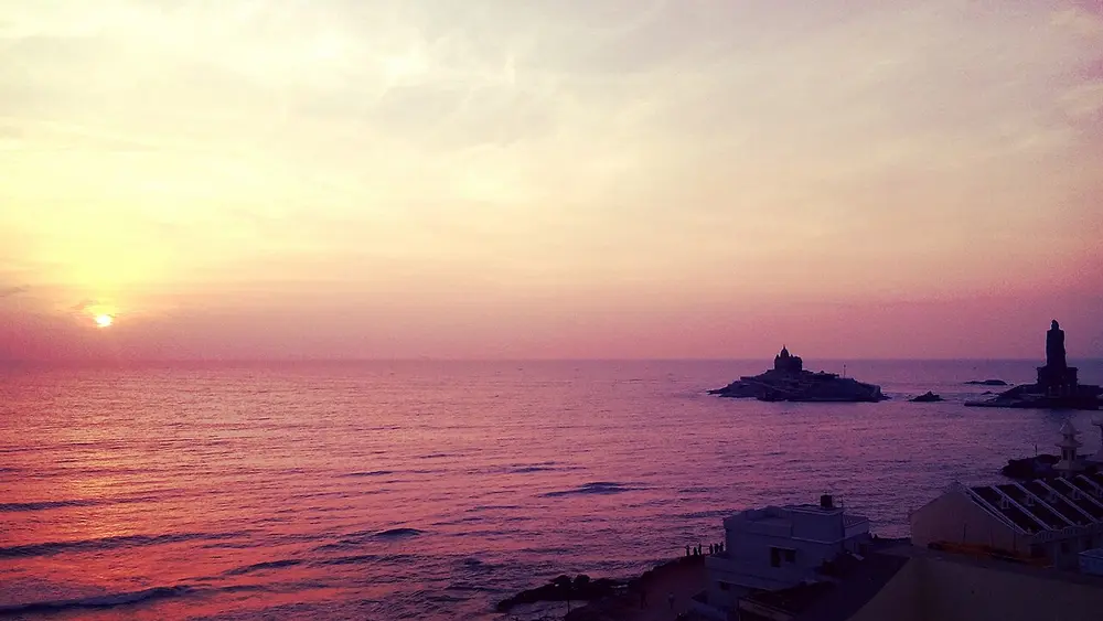 The sun rising over a pink and purple sea, with a small rocky island and a memorial building on it near the shore, and a tall statue on a second rock.