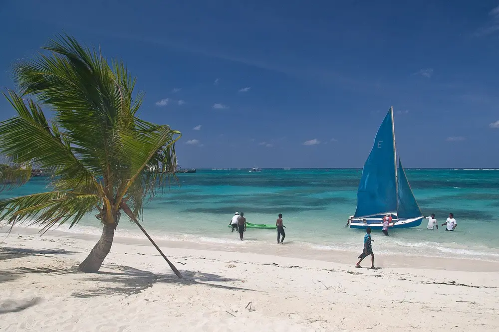 A white sandy beach with a leaning palm tree, turquoise water and a small sailing boat.