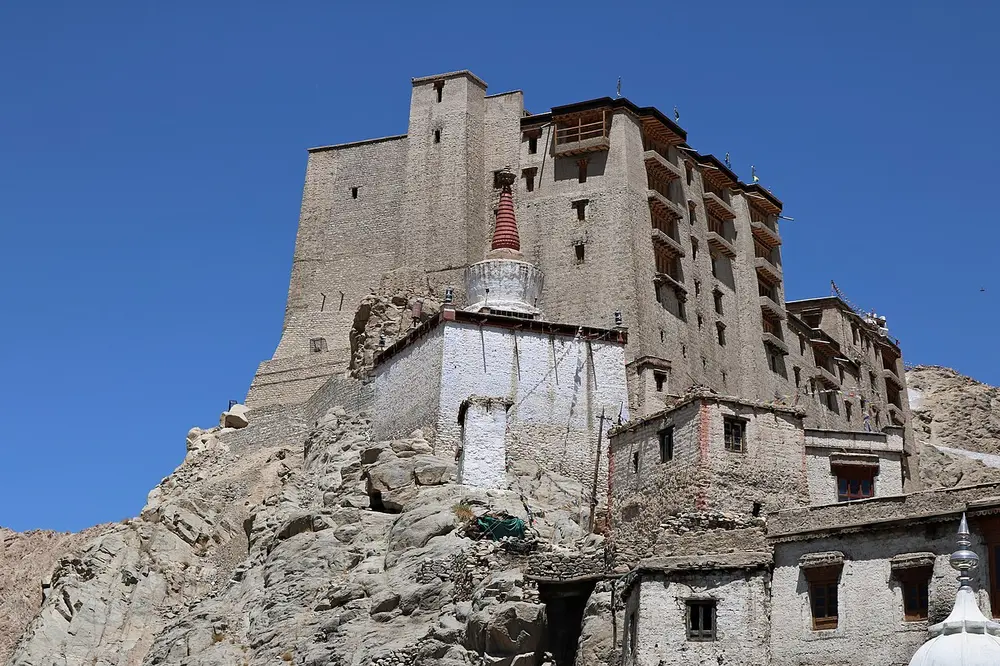 A tall, many-storeyed palace of mud brick and stone on a rocky hill, under a deep blue sky.