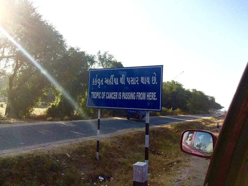 A blue roadside sign in Gujarati and English saying the Tropic of Cancer passes here, beside a tree-lined road.