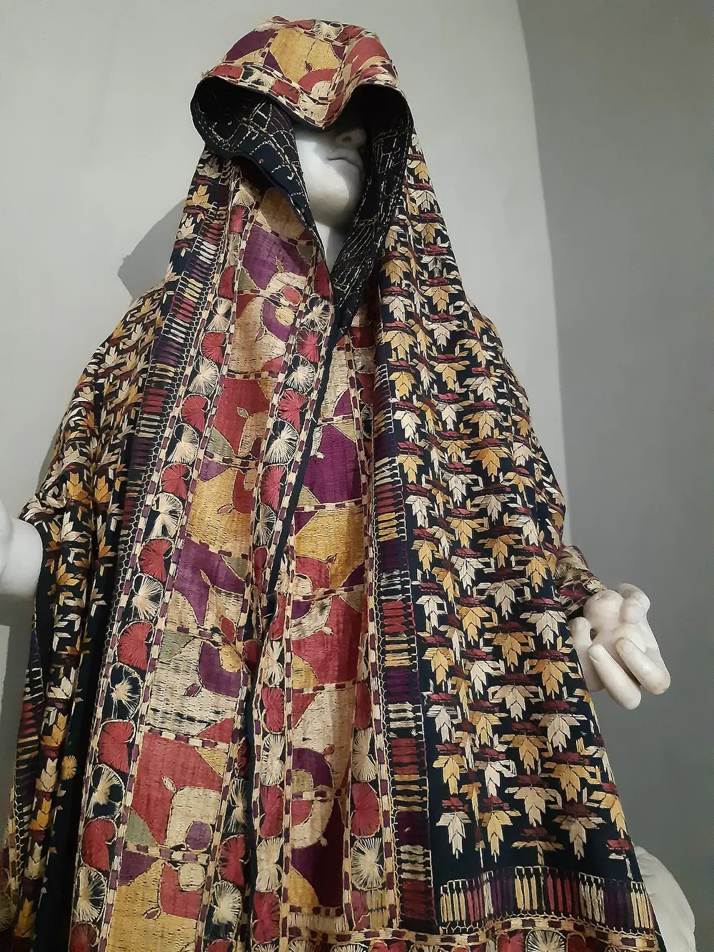 A shawl on a museum figure so densely embroidered in gold, cream, red and purple silk that the dark base cloth barely shows.
