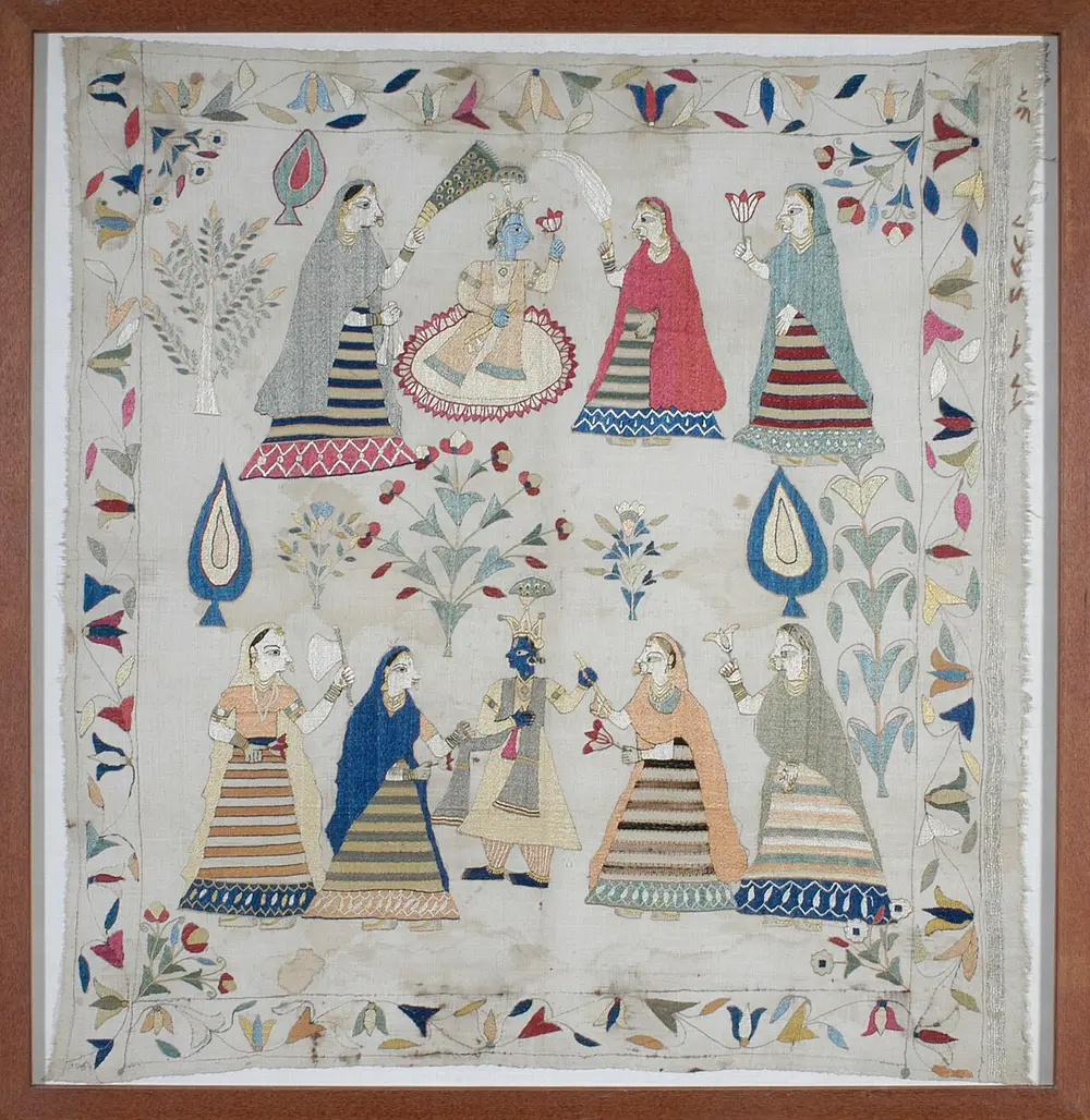 A square embroidered cloth showing Krishna, blue-skinned, with women in striped skirts, between flowering trees and a border of flowers.