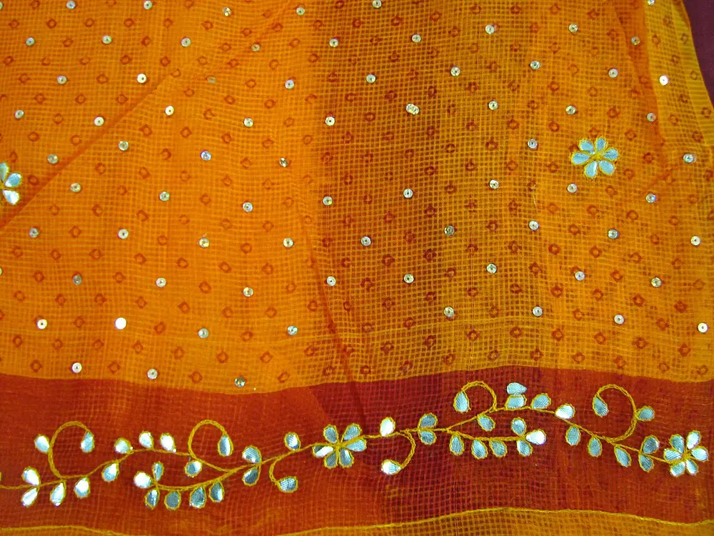 An orange sari with a border of small leaf and flower shapes cut from shiny silver ribbon and stitched on along a curling stem.