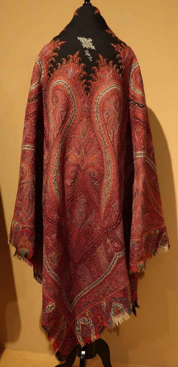 A large red Kashmir shawl on a stand, covered with closely embroidered paisleys and leaves, with a black centre.