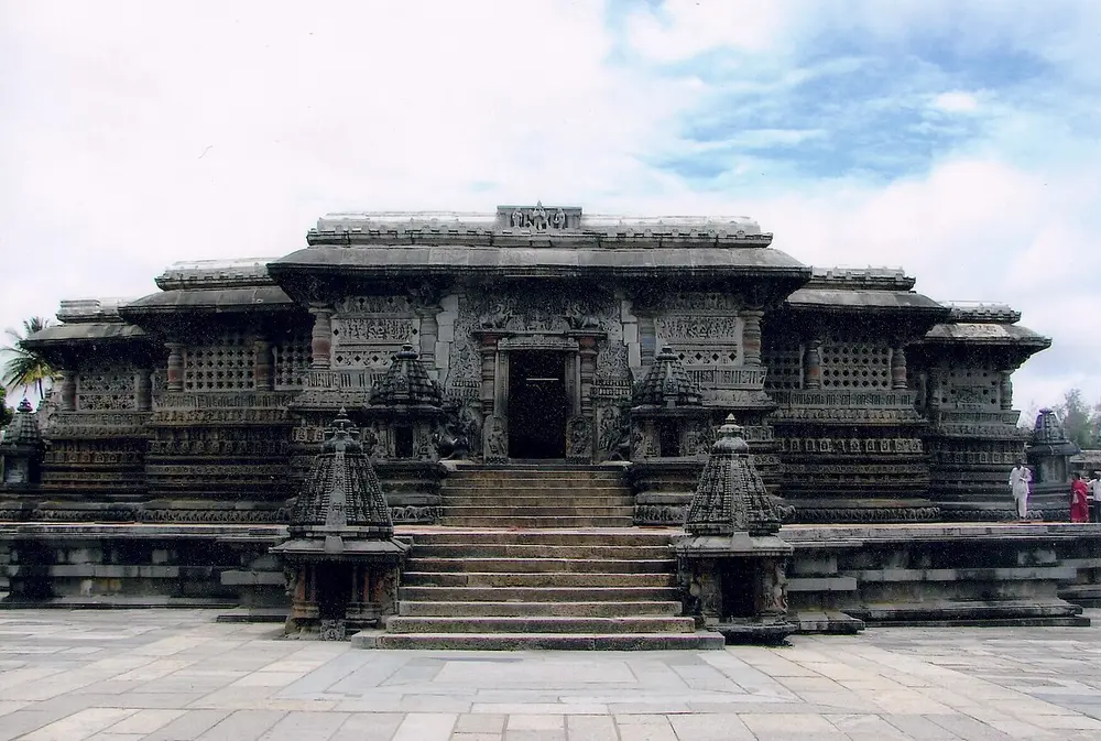 The front of the Chennakeshava Temple at Belur, a low dark stone temple on a raised platform, with a flight of steps flanked by two small shrines and stone screens between its pillars.