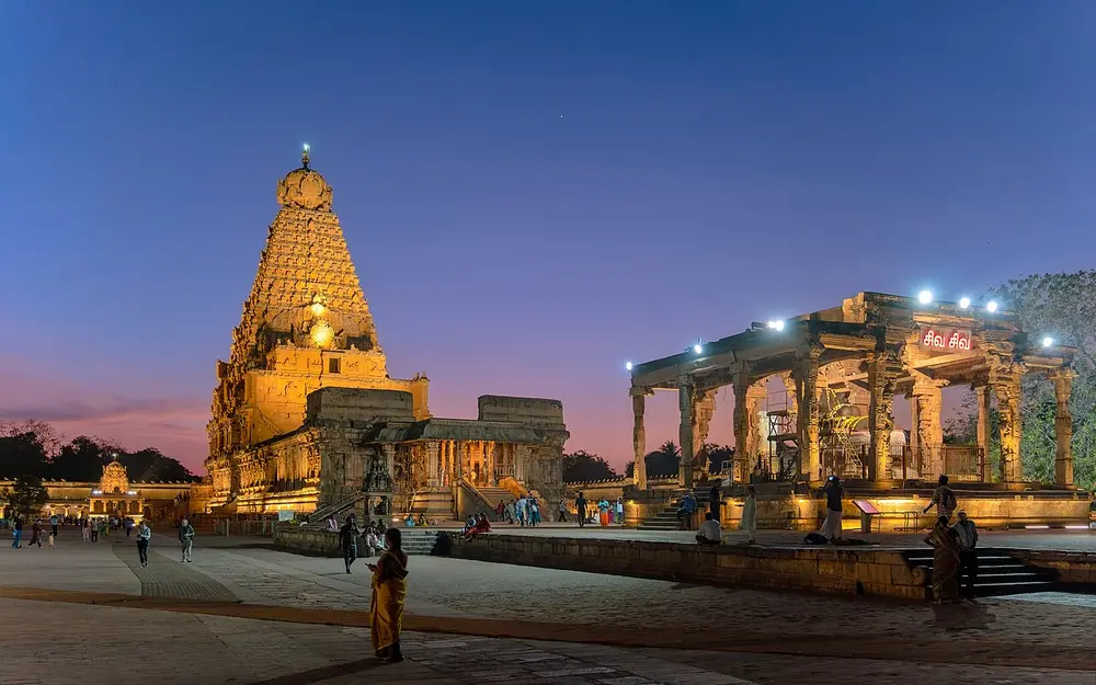 The Brihadeeswara Temple at dusk, its tall granite pyramid tower lit gold, with a pillared Nandi pavilion on the right.