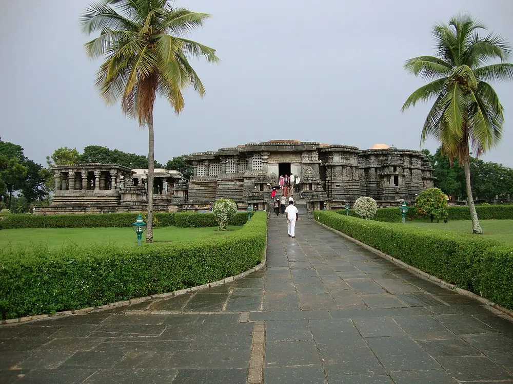 A stone path between hedges and coconut palms leading to the Hoysaleswara Temple at Halebidu, a long, low, flat-roofed stone temple, with a small pillared pavilion on the left.