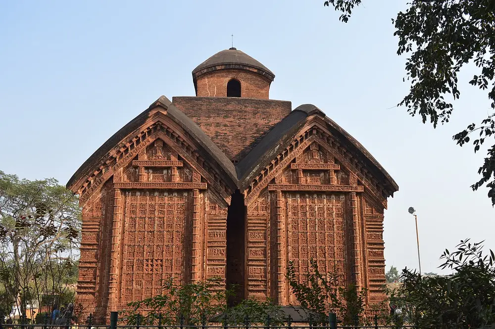 The Jor Bangla Temple at Bishnupur, a red brick temple made of two joined buildings with curved, hut-like roofs, topped by a small tower, its walls covered in carved terracotta panels.