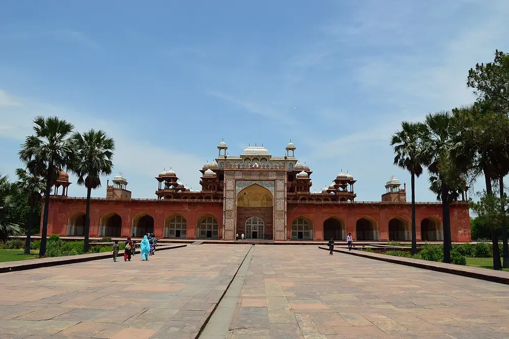 A long red sandstone building with a row of arches and a tall central arched doorway, topped by a small white marble pavilion, seen across a wide paved courtyard with palm trees.