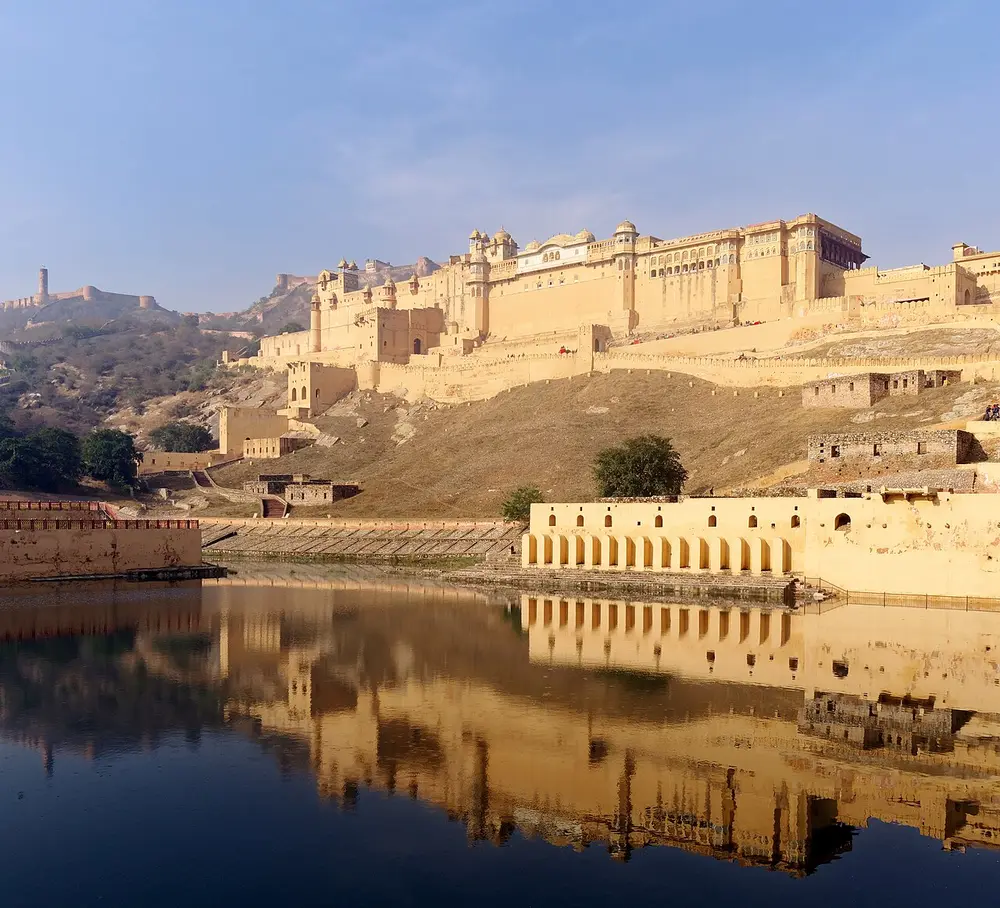 A long cream and yellow palace-fort climbing a hillside in many levels, with walls along the ridges, reflected in a still lake below.