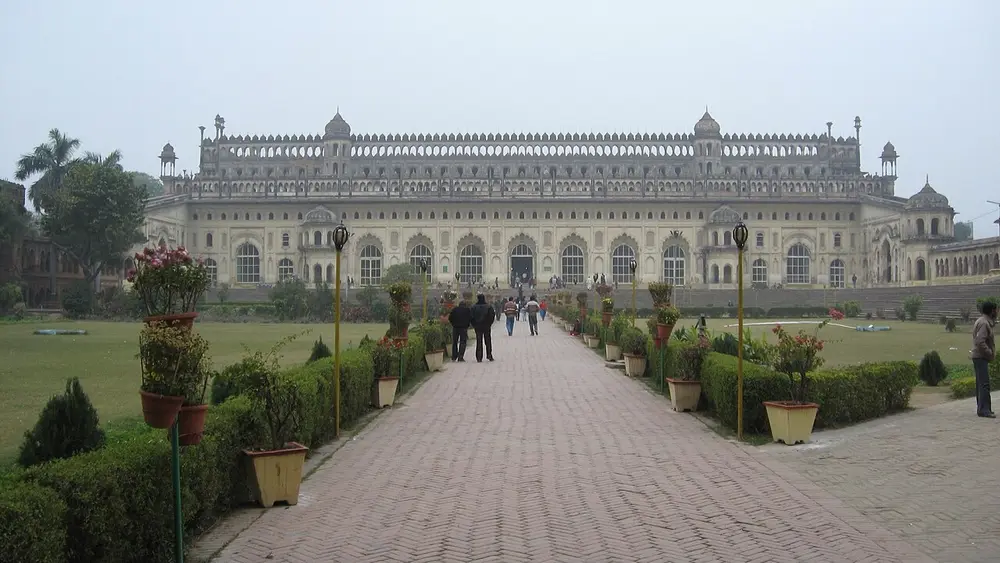 A long, pale two-storey hall with rows of arches and small domed turrets along its roof, at the end of a paved garden path lined with plants.