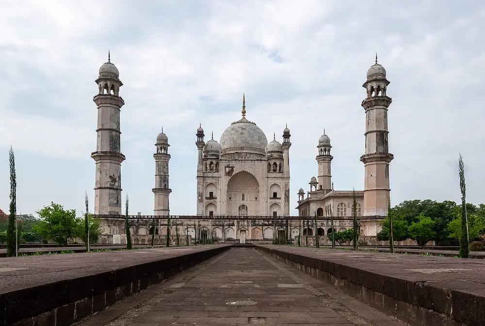A white domed tomb with four tall minarets at the corners of its platform, seen at the end of a long raised walkway with a water channel.