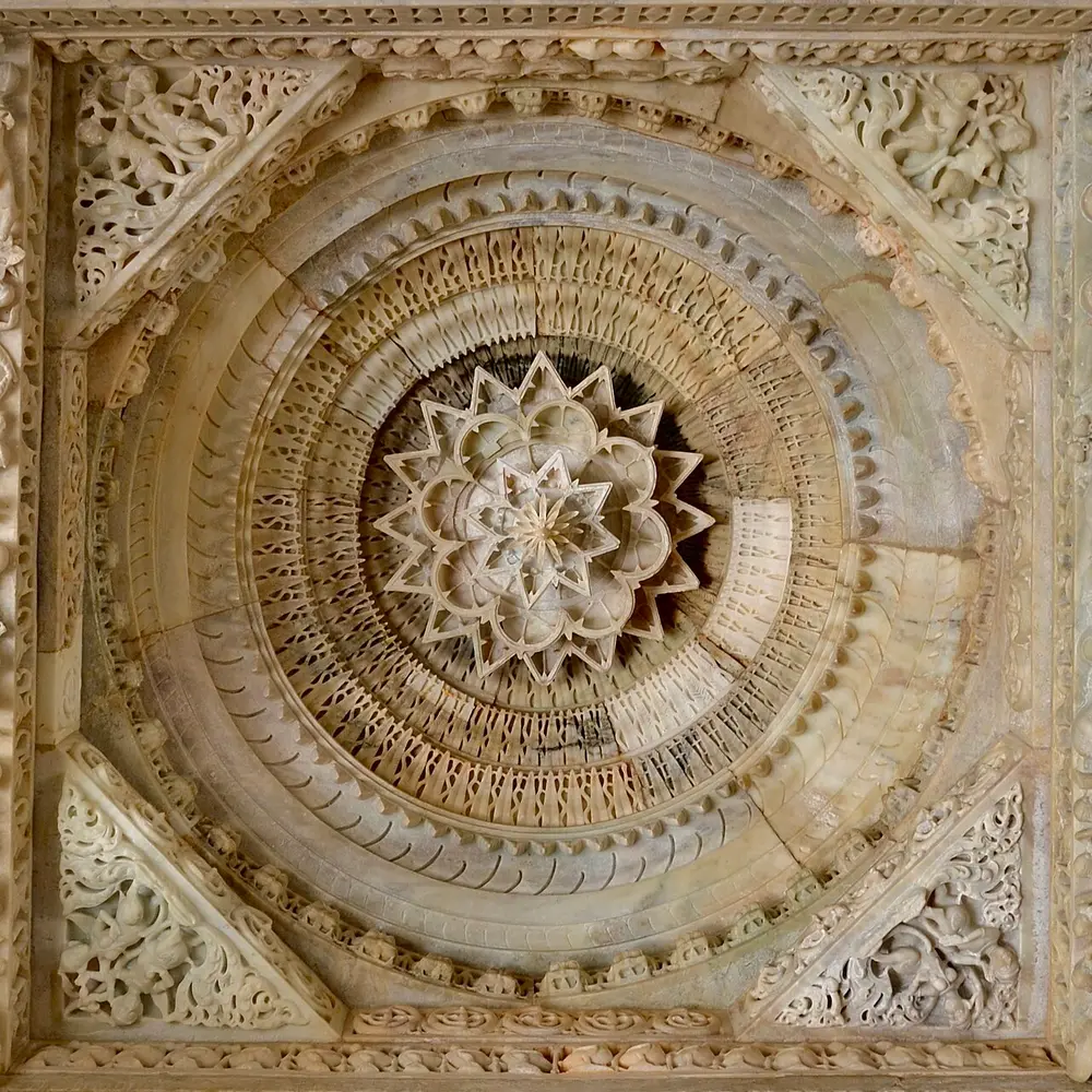 A round white marble ceiling carved in rings of fine lace-like patterns around a hanging lotus at the centre, inside a square frame with carved corners.