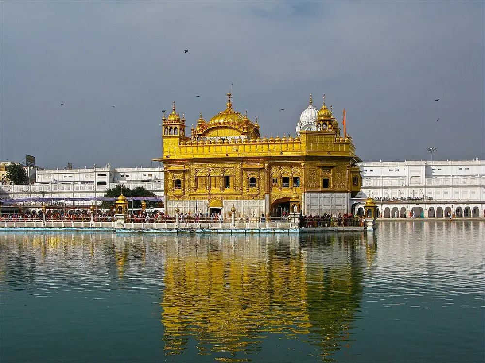 A two-storey shrine covered in gold, with a gold dome, standing in a large still pool that reflects it, with long white buildings around the edge of the pool.