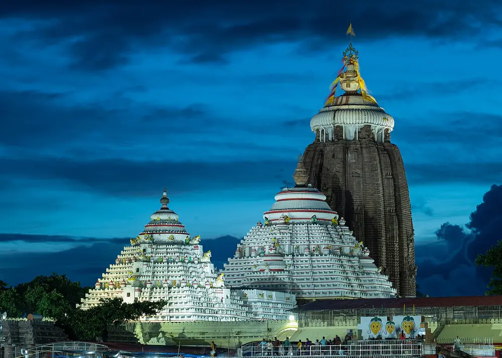 The tall curved stone tower of the Jagannath Temple rising above smaller white halls with stepped pyramid roofs, lit up against a dark blue evening sky.