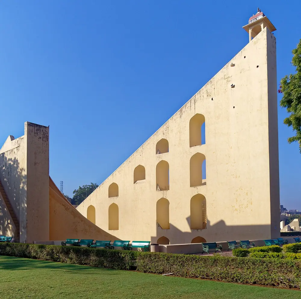A huge cream-coloured stone triangle with arched openings in its side and a stair along its sloping edge, beside a curved stone scale, on a green lawn.