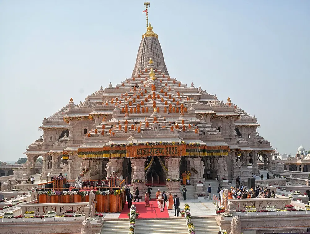 A large pale sandstone temple with a tall curved central spire and many smaller spires, hung with orange marigold garlands, with people on the steps in front.
