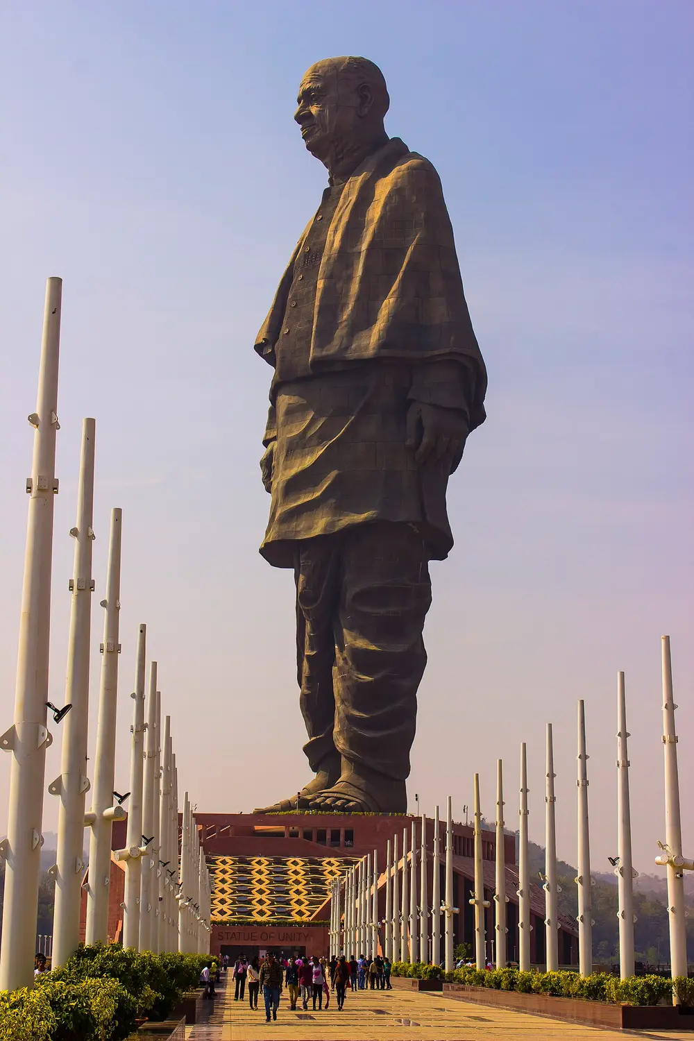 A colossal dark bronze statue of Sardar Patel in a long shawl, standing on a tall base at the end of an avenue of tall white poles.