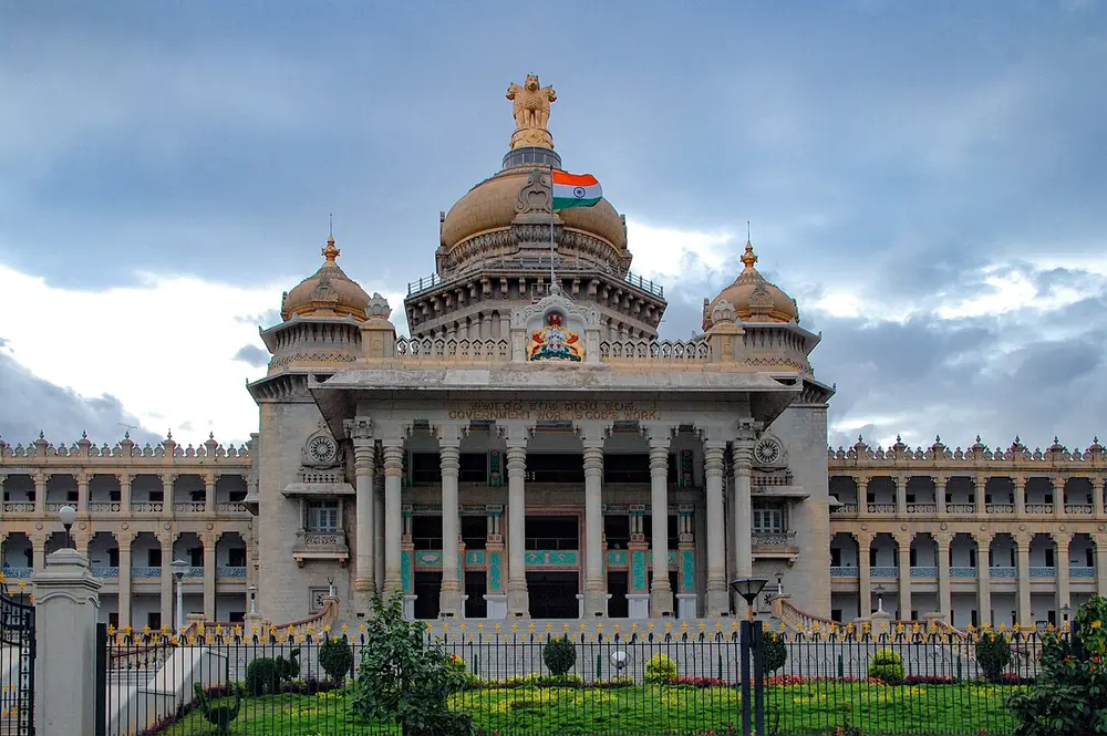 A huge grey stone government building with a broad flight of steps, a row of tall columns, a large central dome topped by the four-lion capital, and smaller domes, under a cloudy sky.