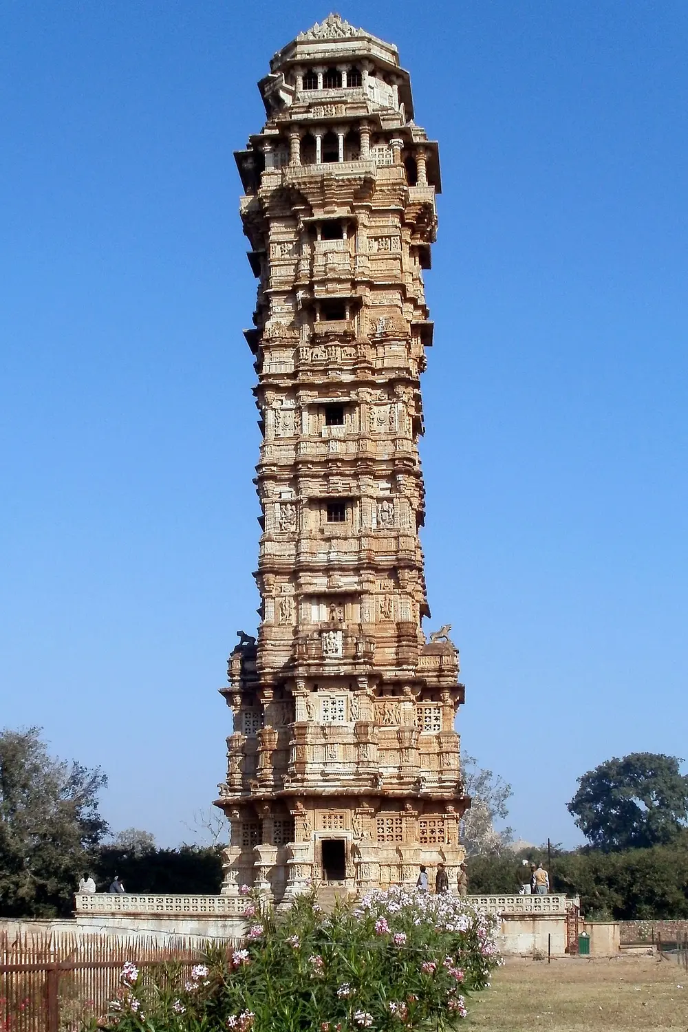 A tall, slim stone tower of nine storeys covered in carved figures, with small balconies and a domed top, against a clear blue sky.