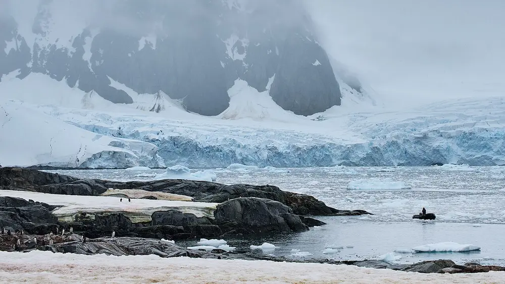 A wall of blue-white glacier ice below dark, misty mountains, with rocks, floating ice and a few penguins in the foreground.
