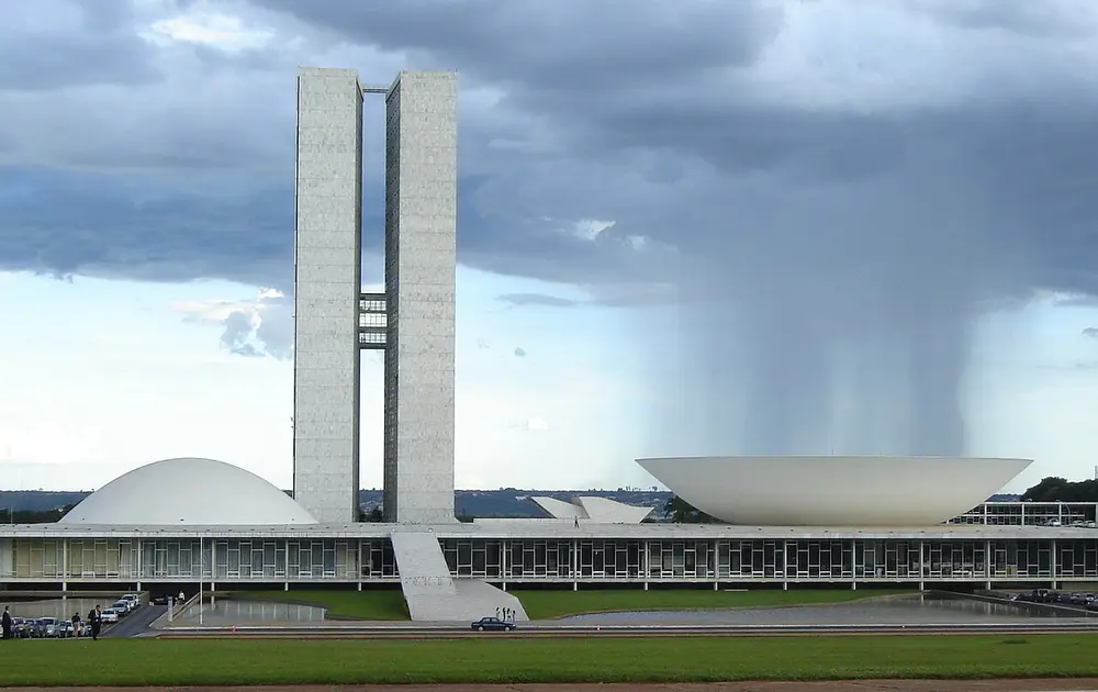 A modernist government complex with two tall slim towers, a white dome on one side and a white upturned bowl on the other, under dark rain clouds.
