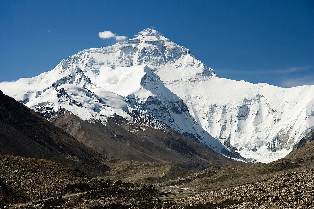 A huge snow-covered pyramid of a mountain under a deep blue sky, rising above a brown, rocky valley.
