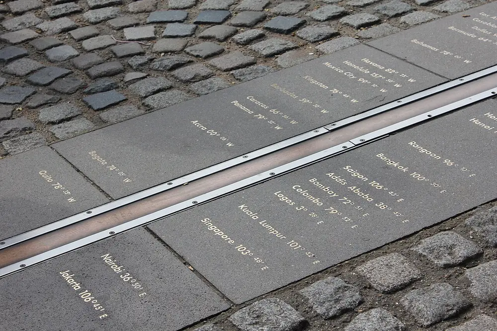 A metal strip set into paving stones, with the names of world cities and their longitudes written on either side.