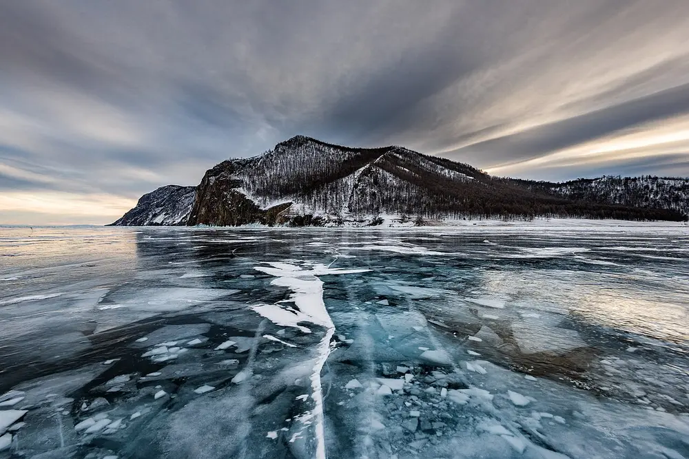 A frozen lake with clear, cracked blue ice in the foreground and a dark forested hill beyond, under streaky grey clouds.