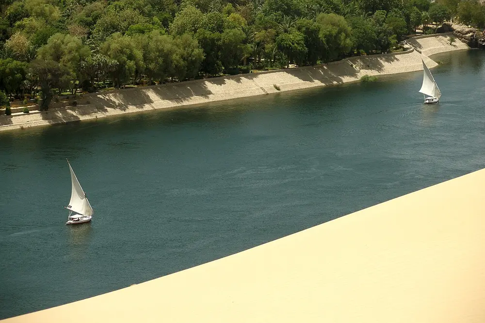 Two white-sailed wooden boats on a wide blue river, with a green bank of trees on the far side and pale desert sand in the foreground.