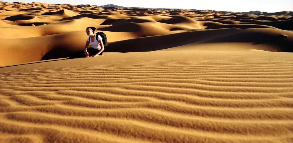 Rows of golden sand dunes with fine ripples in the sand, and a person in a white T-shirt sitting on a dune.