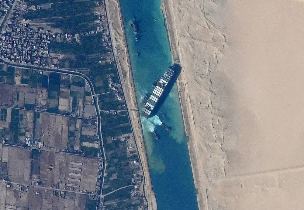 A photo from space of a straight canal through the desert, with a long container ship stuck diagonally across it.
