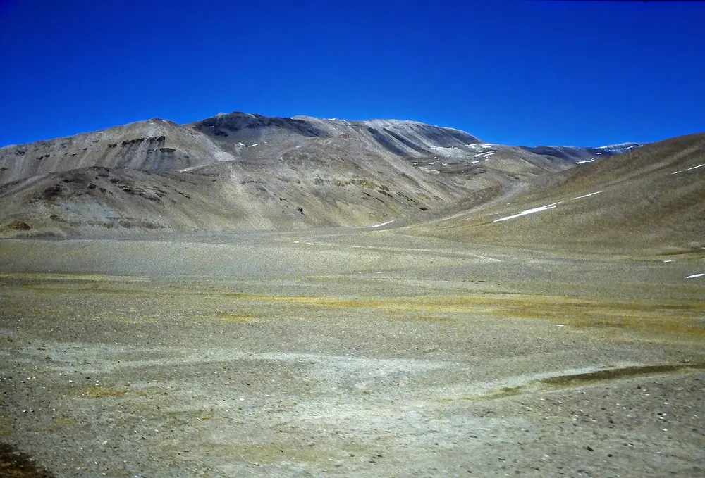 A wide, bare, gravelly plain at high altitude with rounded brown hills and patches of snow under a dark blue sky.