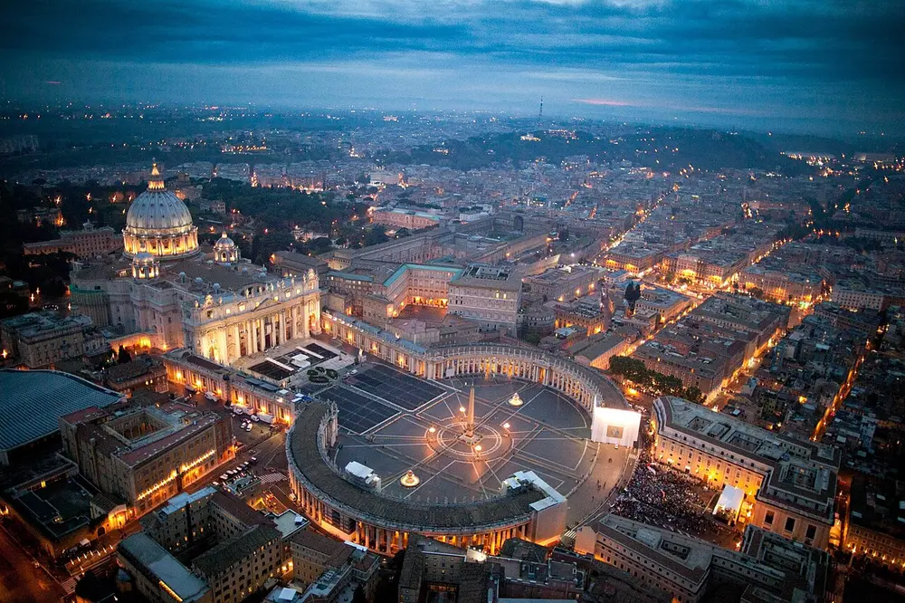 An aerial view at dusk of a huge domed church facing a vast oval square ringed by curved rows of columns, with city lights all around.