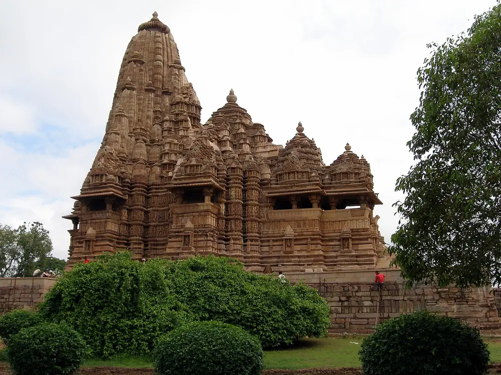 A tall sandstone temple whose roofline rises in a cluster of curved peaks, behind green bushes.