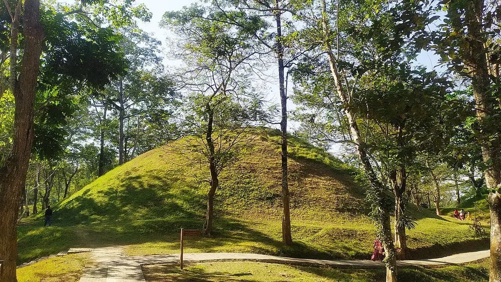 A large grassy mound shaped like a small hill, among tall trees, with a paved path in front.