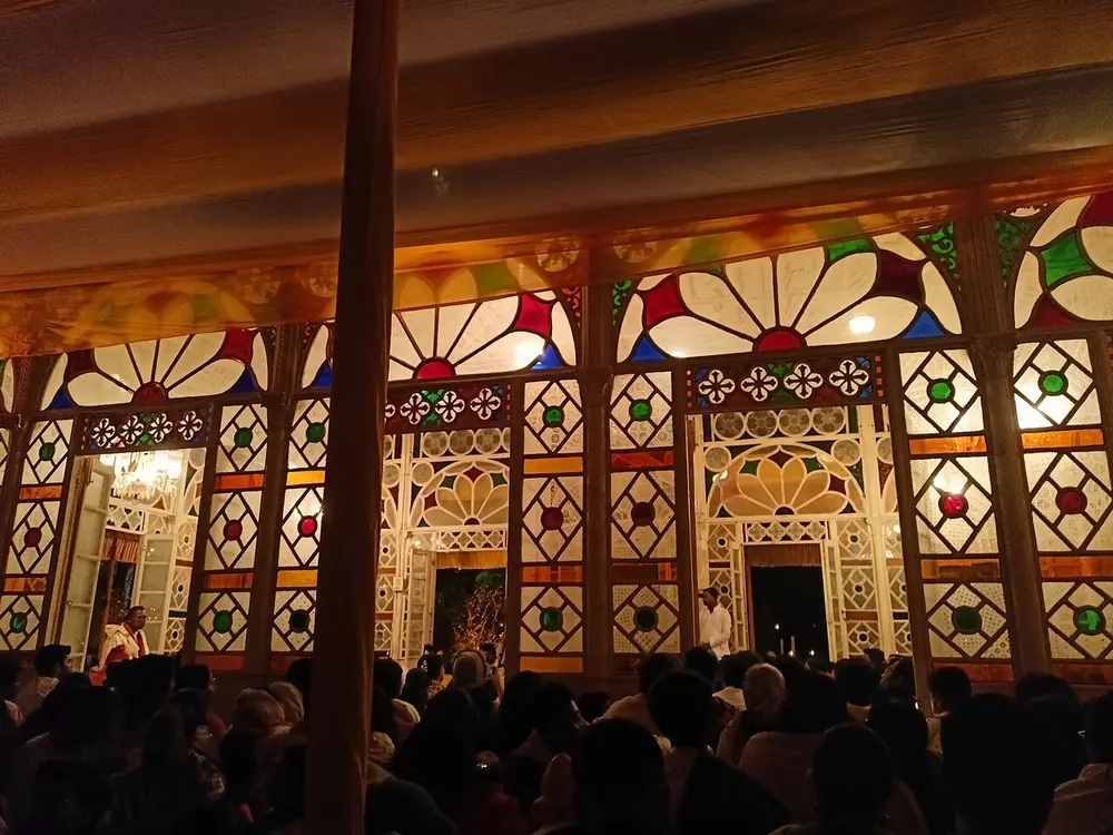 A prayer hall at night with walls of coloured glass in flower patterns, and people seated in front.