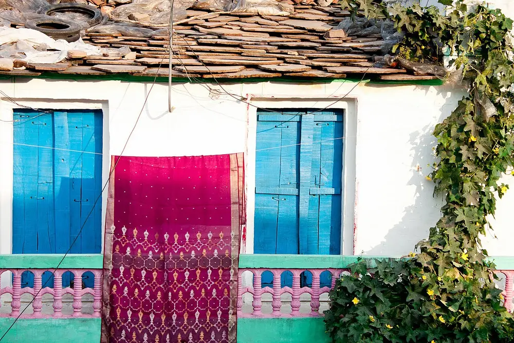 A sheer magenta sari hung over a balcony railing between two blue windows, with small gold and white motifs woven along its lower part.