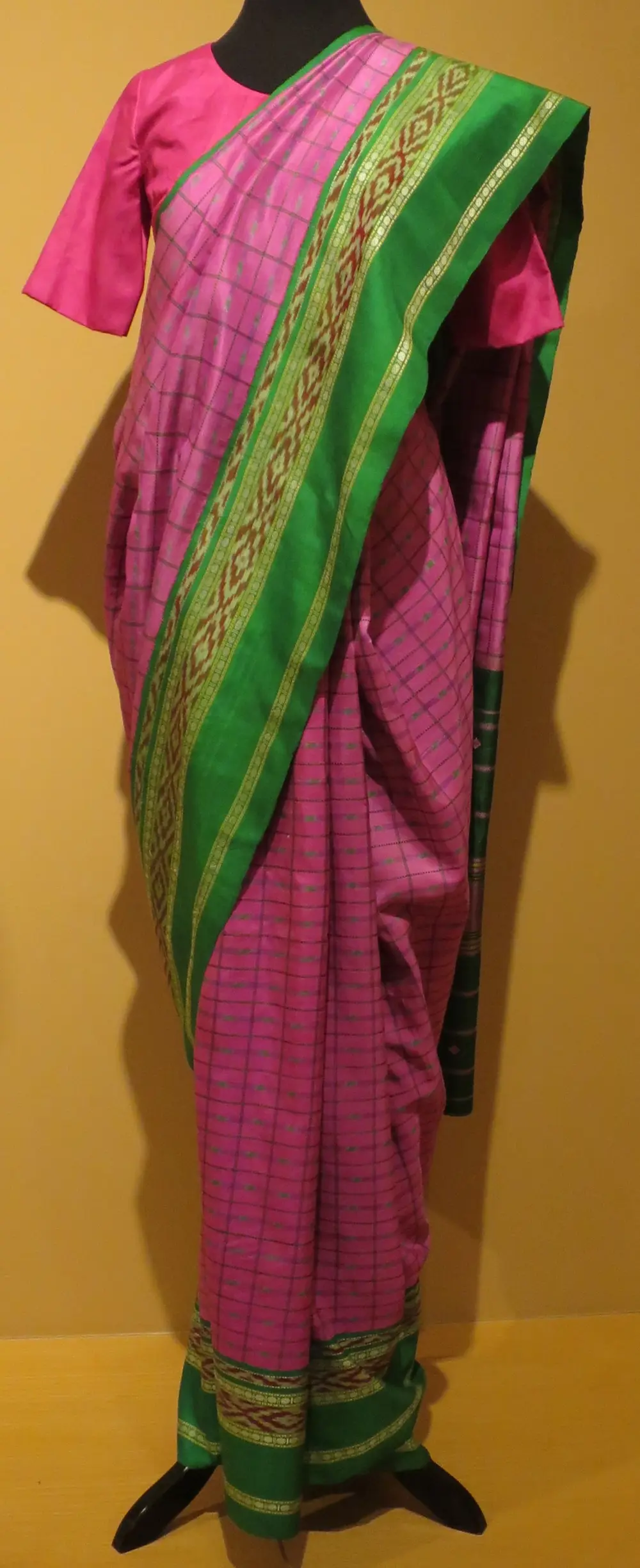 A pink silk sari with a fine check pattern, draped on a stand, with a wide bright green border and pallu edged in gold.