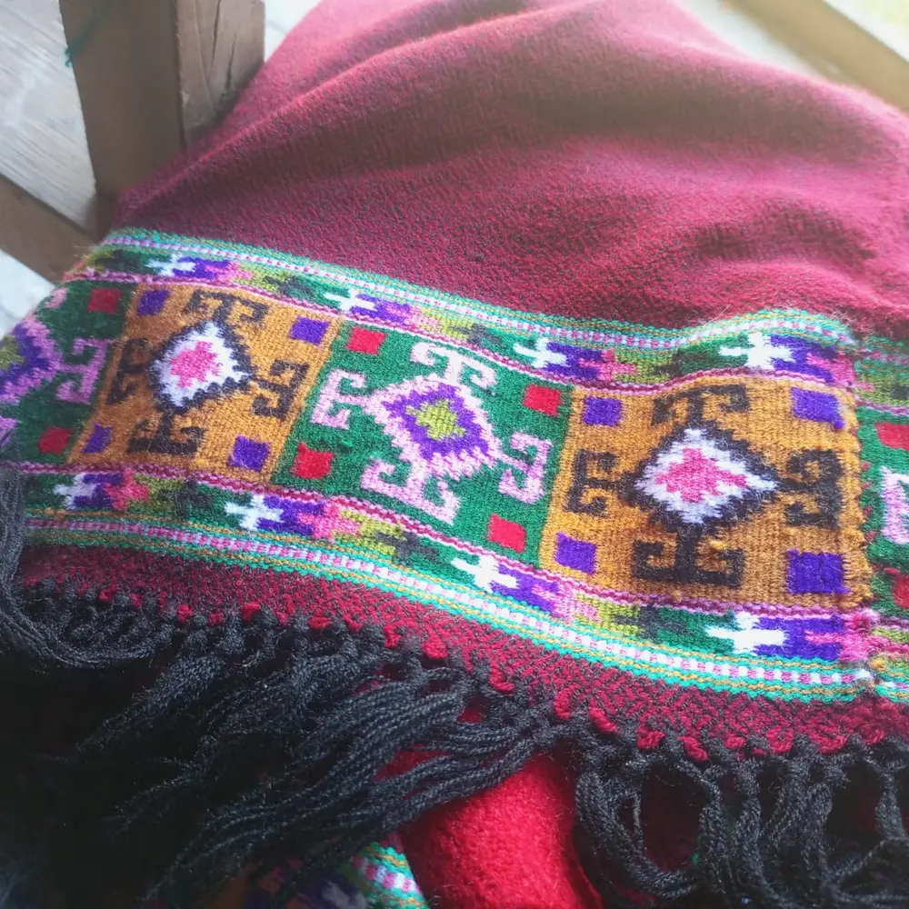 A maroon woollen Kullu wrap with black fringes and a wide woven border of geometric shapes in mustard, green, pink and purple.
