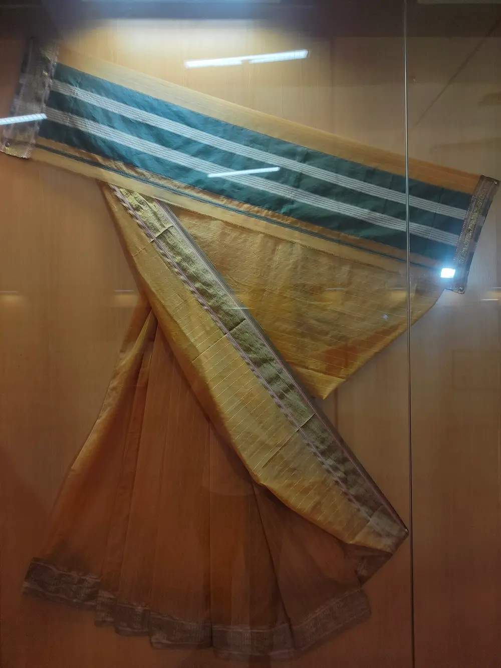 A pale gold sari in a museum case, with a green pallu crossed by white stripes and narrow patterned borders.