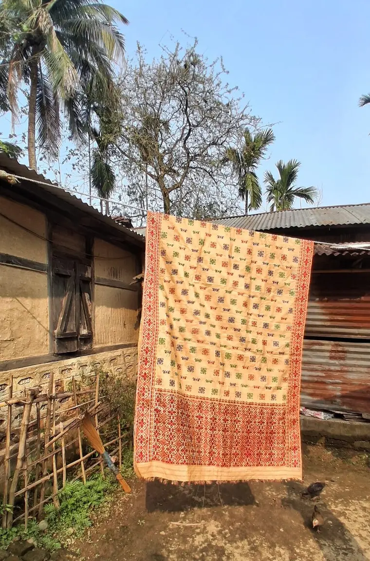 A golden-cream silk mekhela chador hung outside a village house, woven with small red, green and blue motifs and a deep red patterned border.