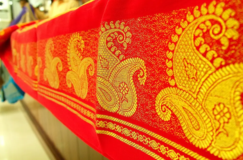 A red silk sari border with large gold zari paisleys, laid over a display counter.